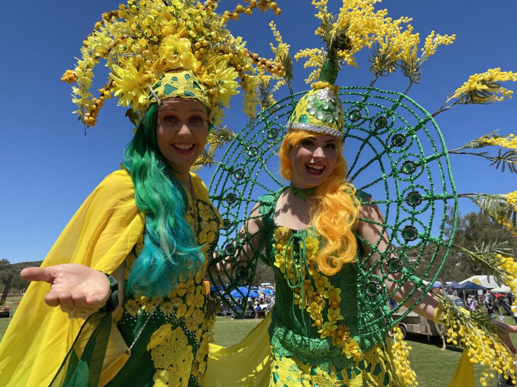 Two stunning stilt walkers dressed in green and yellow against the backdrop of a bright blue sky.